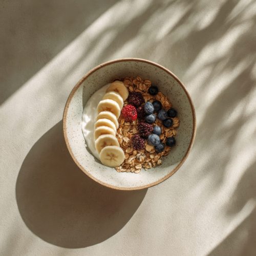 Healthy breakfast bowl with oatmeal, yogurt, banana slices, blueberries, and raspberries under harsh natural sunlight